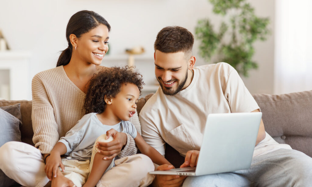 Diverse family using laptop on sofa together Happy multiethnic parents and amazed mixed race boy watching video on laptop while resting on couch at home together
